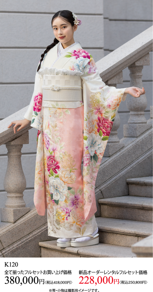 白とピンクの花柄の着物を着た女性が屋外に立ち、穏やかな笑顔でカメラを見つめている。黒髪をヘアアクセサリーでスタイリングし、片手を上げてポーズをとっている。着物の価格は以下の通り。.