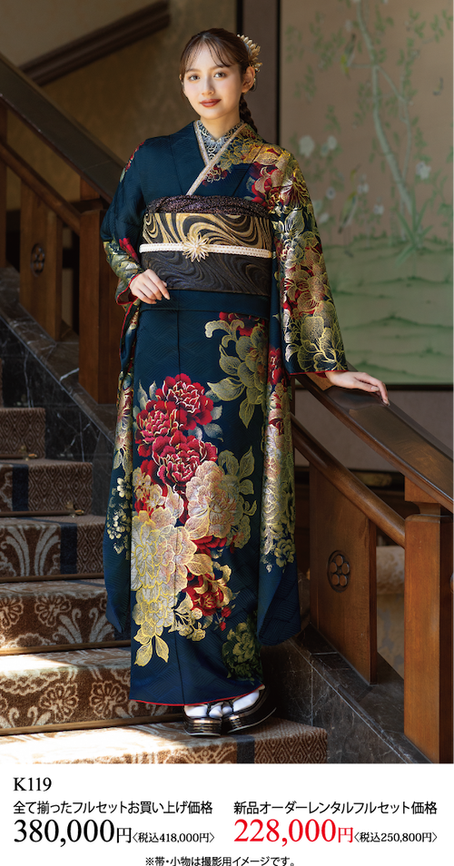 金と赤の模様が入った濃い色の花柄の着物を着た若い女性が屋内に立っている。カーペットを敷いた階段で笑顔でポーズをとっている。着物の購入またはレンタルの価格情報は以下に表示されています。.